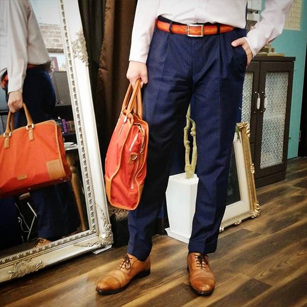 紺のスラックスに茶色の革靴とベルトを合わせた男性が、革のバッグを2つ持っている