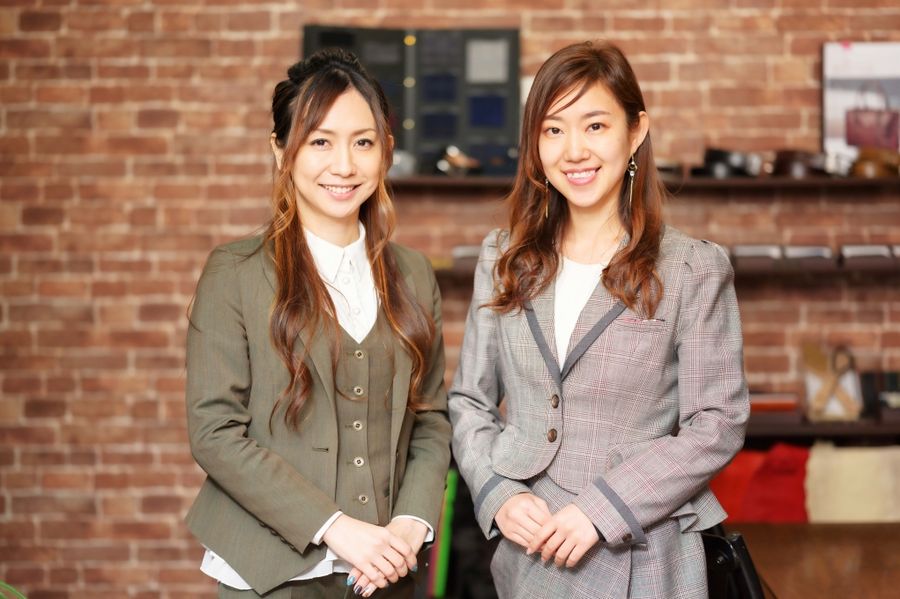 スーツを着た女性2人が、スーツの並ぶ店内のレンガ壁の前に並んで立っている